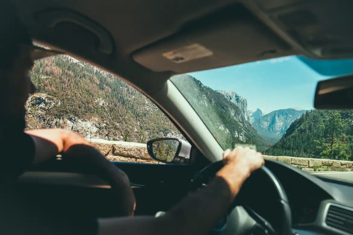 Une Personne Conduit Une Voiture Dans Un Paysage De Montagne Pittoresque, Visible à Travers Le Pare Brise, Avec Des Arbres Et Des Falaises Rocheuses Au Loin, Sous Un Ciel Dégagé.
