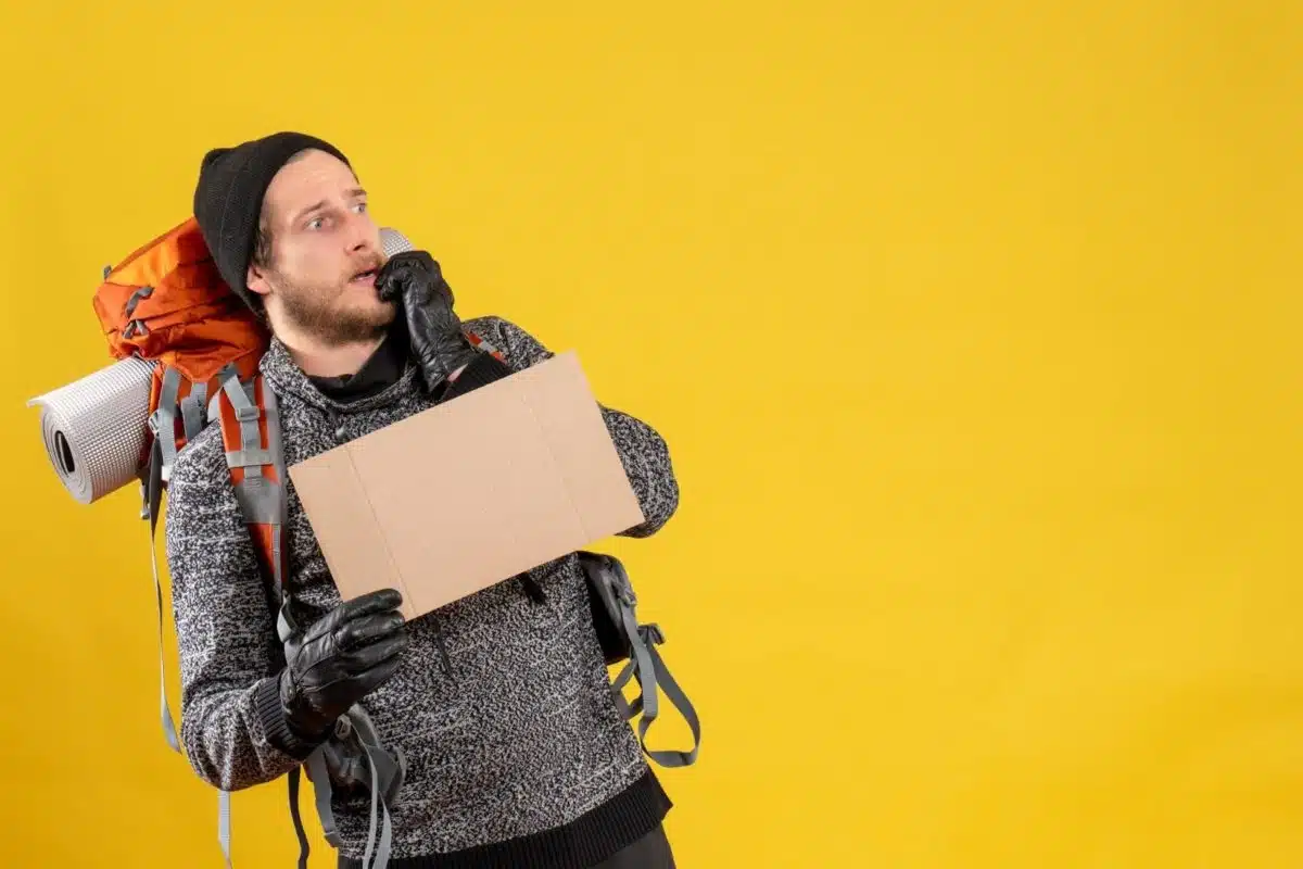 Un Homme Portant Un Sac à Dos Et Des Gants Tient Une Pancarte En Carton Vierge Et Semble Préoccupé Sur Un Fond Jaune.