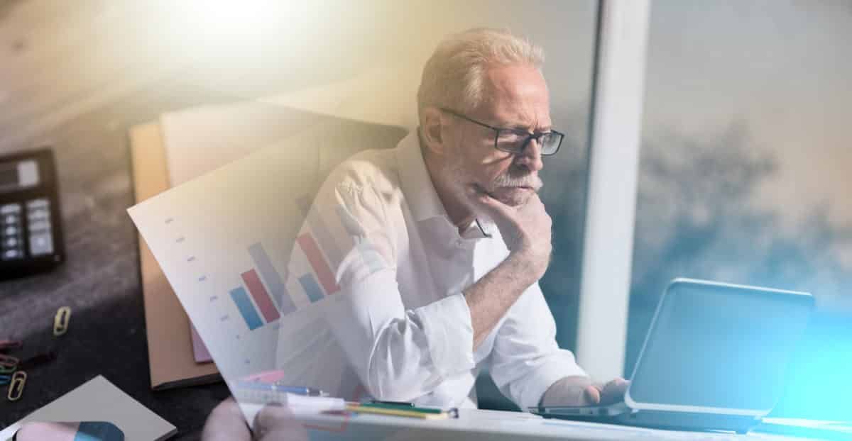 Un Homme D'âge Mûr Avec Des Lunettes Regarde Pensivement Un Ordinateur Portable, Avec Des Tableaux Et Des Fournitures De Bureau Visibles, Ce Qui Suggère Un Cadre De Travail Ou D'analyse.