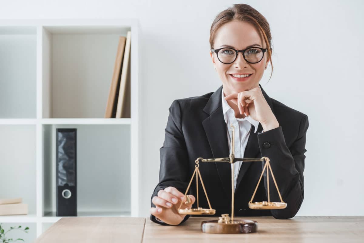 Une Femme En Tailleur Est Assise à Un Bureau Avec Une échelle De Justice Et Sourit à La Caméra. Des étagères Contenant Des Livres Et Des Dossiers Se Trouvent à L'arrière Plan.