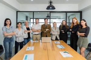 Un Groupe De Neuf Professionnels Se Tient Debout, Les Bras Croisés, Derrière Une Table De Conférence équipée De Tableaux Et D'un Ordinateur Portable, Dans Une Salle De Réunion Moderne.