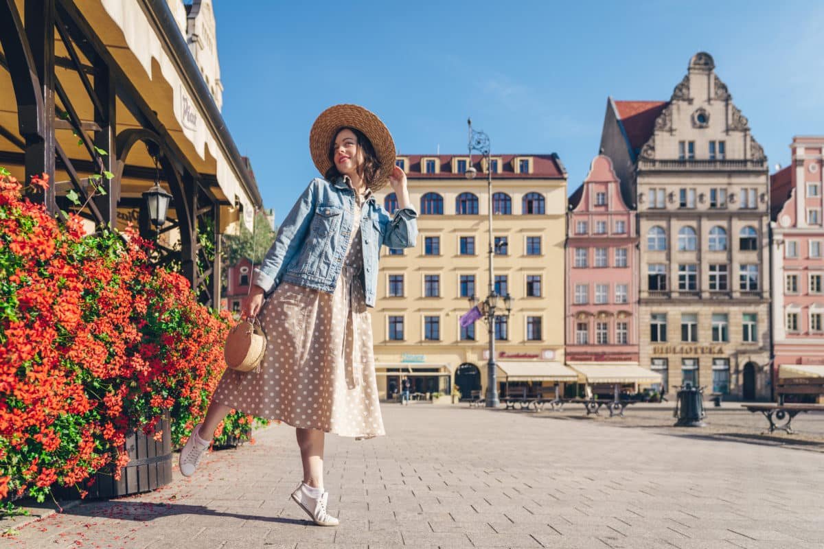 Une Femme En Robe à Pois Et Chapeau De Paille Pose Près De Fleurs Rouges Sur Une Place Ensoleillée De La Ville Avec Des Bâtiments Historiques En Arrière Plan.