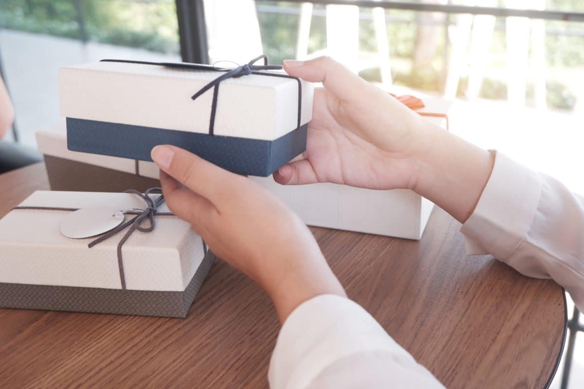 Une Personne Tenant Un Paquet Cadeau Emballé Avec Un Ruban Bleu à Une Table, Avec D'autres Paquets Cadeaux En Arrière Plan.