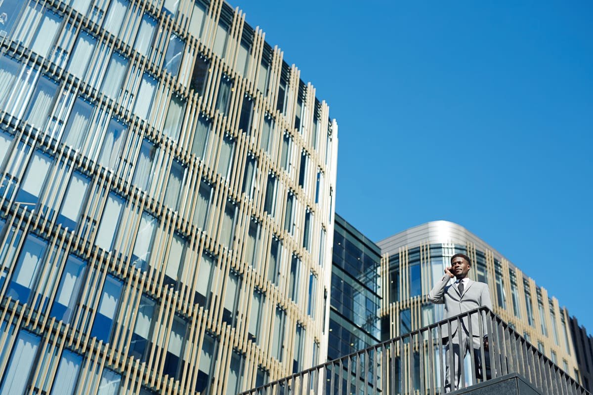 Un Homme En Costume Se Tient Sur Un Escalier Extérieur Et Parle Au Téléphone, Avec En Arrière Plan Des Immeubles De Bureaux Modernes En Verre Et Un Ciel Bleu Clair.