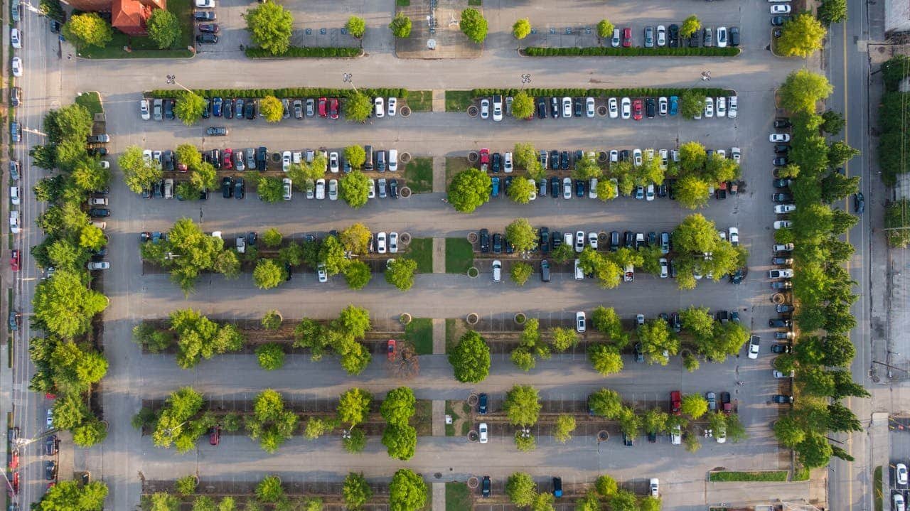 Vue Aérienne D'un Parking Avec Plusieurs Rangées De Voitures Garées Et Des Arbres Verts Régulièrement Espacés.