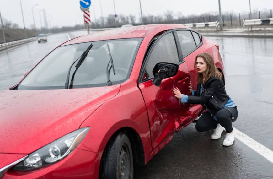 Une Femme Est Agenouillée à Côté D'une Voiture Rouge Dont La Portière Est Endommagée Et Le Rétroviseur Latéral Manquant, Sur Une Route Mouillée.