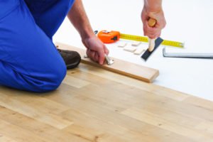 Personne Installant Un Parquet, Utilisant Un Marteau Et Une Cale Pour Assembler Les Planches ; Un Ruban De Mesure Et Des Cales Sont Visibles Sur Le Sol.