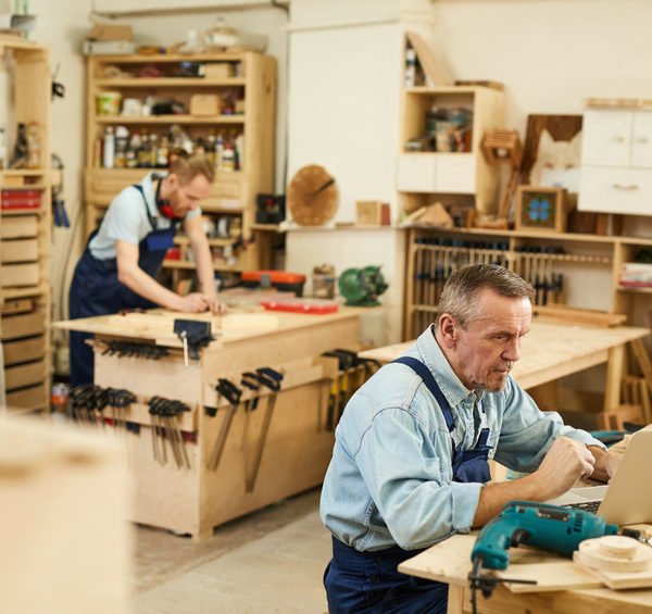 Deux Hommes Dans Un Atelier De Menuiserie ; L'un Travaillant Sur Un Ordinateur Portable à Une Table Au Premier Plan, L'autre Mesurant Du Bois à Un établi à L'arrière Plan.