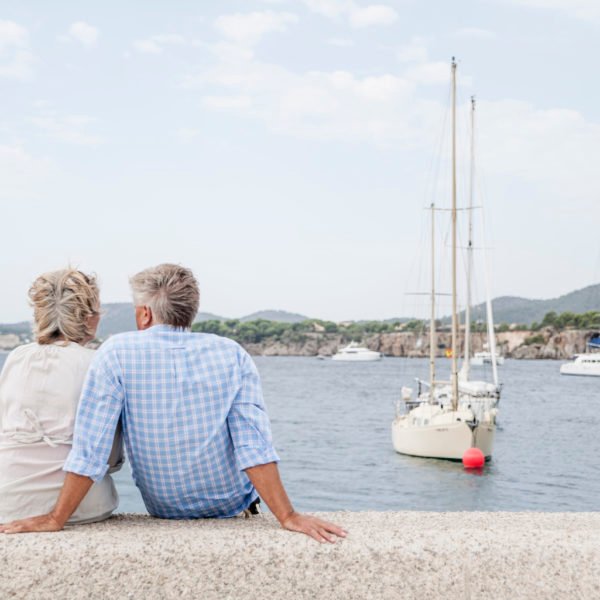 Un Couple âgé Est Assis Sur Une Corniche En Pierre Au Bord De La Mer, Regardant Les Voiliers Ancrés Dans L'eau Et Les Collines Visibles à L'arrière Plan.
