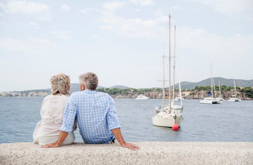 Un Couple âgé Est Assis Sur Une Corniche En Pierre Au Bord De La Mer, Regardant Les Voiliers Ancrés Dans L'eau Et Les Collines Visibles à L'arrière Plan.