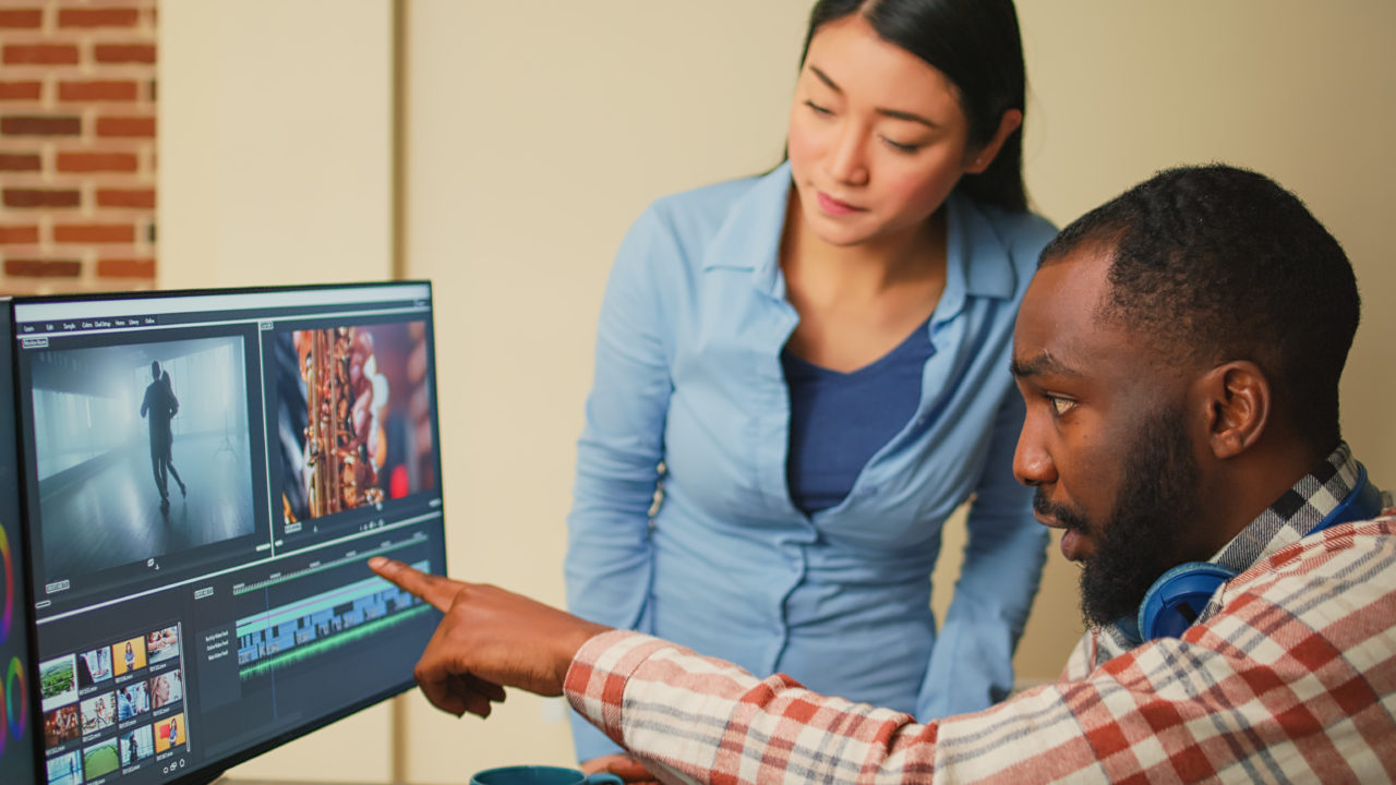 Un Homme Montre Une Ligne De Temps De Montage Vidéo Sur Un écran D'ordinateur Tandis Qu'une Femme Se Tient à Côté De Lui, Tous Deux Concentrés Sur L'écran.