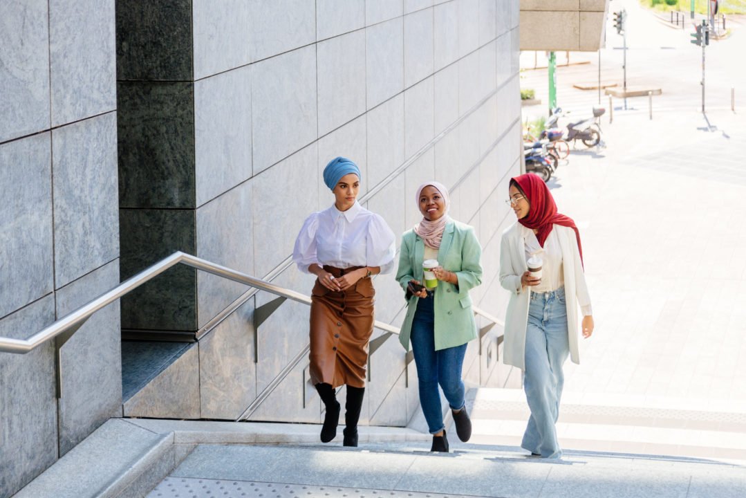 Trois Femmes Portant Un Foulard Montent Des Escaliers Extérieurs à Côté D'un Bâtiment Moderne, En Discutant Et En Souriant ; L'une D'elles Tient Une Tasse De Café.