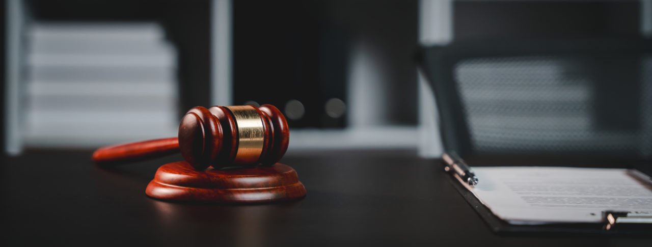 Un Marteau De Juge En Bois Repose Sur Un Bloc Sonore Sur Un Bureau Sombre, Avec Un Presse Papiers Et Une Pile De Livres à L'arrière Plan.