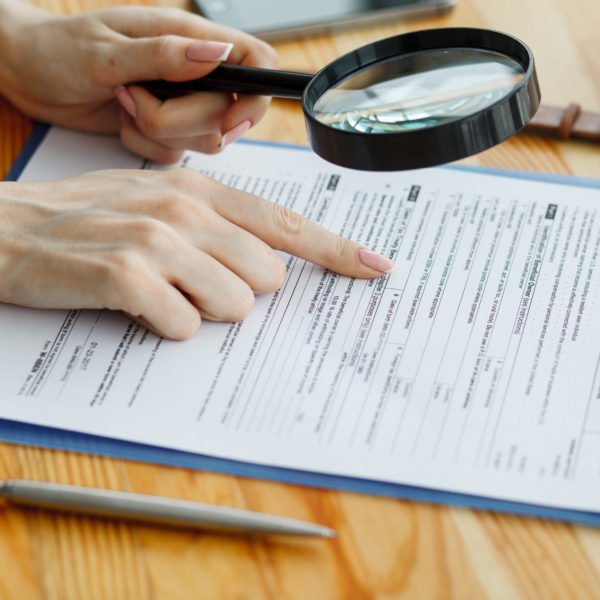 Une Personne Tient Une Loupe Et Pointe Une Partie D'un Formulaire Sur Un Presse Papiers, Avec Un Stylo Et Un Smartphone Sur Un Bureau En Bois.