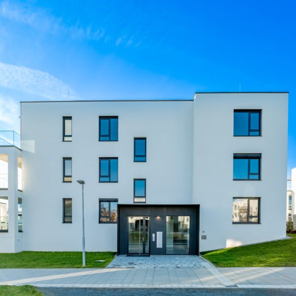 Immeuble Moderne De Trois étages Avec Un Extérieur Blanc, De Grandes Fenêtres Et Une Pelouse Sous Un Ciel Bleu Clair.