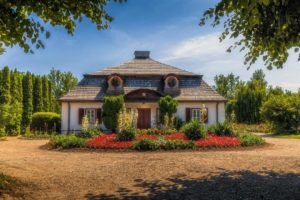 Une Petite Maison Pittoresque D'un Seul étage Avec Un Toit En Bois, Des Fenêtres Rondes Et Un Jardin De Fleurs Colorées Devant, Entourée D'arbres Sous Un Ciel Bleu.