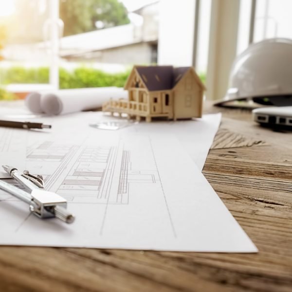 Des Plans D'architecte, Un Compas, Une Maquette De Maison, Des Plans Roulés Et Un Casque Blanc Sont Posés Sur Un Bureau En Bois Près D'une Fenêtre éclairée Par Le Soleil.