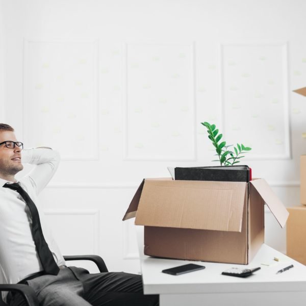 Un Homme En Tenue De Travail Est Assis, Détendu, à Un Bureau Où Se Trouvent Des Cartons De Déménagement Et Une Plante, Semblant S'installer Dans Un Nouveau Bureau.