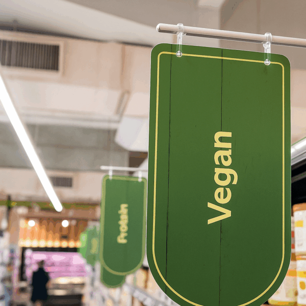 Un Panneau Vert De L'épicerie Portant Le Mot "vegan" Est Suspendu Au Dessus D'une Allée, Indiquant La Section Réservée Aux Produits Végétaliens.