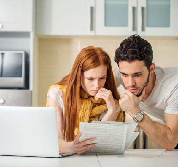 Une Femme Et Un Homme Dans Une Cuisine Regardent Des Documents Avec Des Expressions Préoccupées Tout En Utilisant Un Ordinateur Portable.
