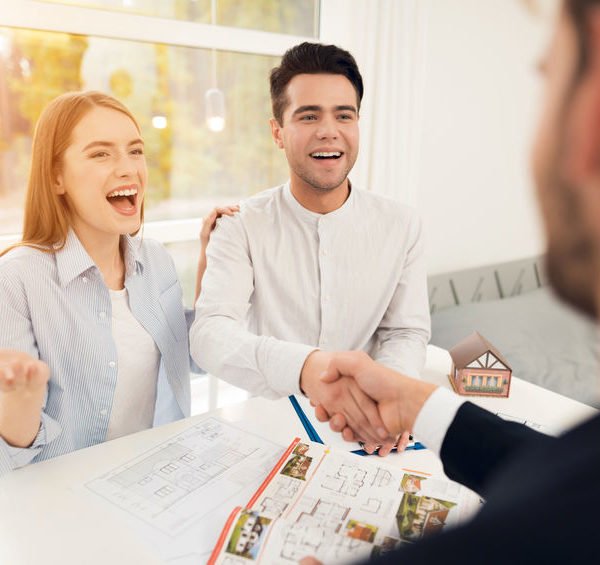 Un Couple Souriant Est Assis à Une Table Avec Des Plans De Maison Et Une Maison Miniature, Tout En Serrant La Main D'une Autre Personne, Ce Qui Suggère Une Réunion Sur L'immobilier Ou L'achat D'une Maison.
