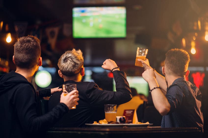 Trois Personnes Sont Assises à Une Table Dans Un Bar, Tenant Des Boissons Et Regardant Un Match De Football Sur Un écran De Télévision En Arrière Plan.