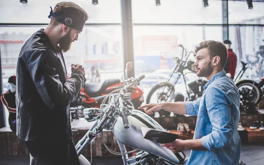 Deux Hommes Inspectent Une Moto Dans Une Salle D'exposition ; L'un Est Debout Et Porte Une Veste En Cuir Et Un Bandeau, L'autre Est Assis, Tient Une Brochure Et Montre La Moto Du Doigt.