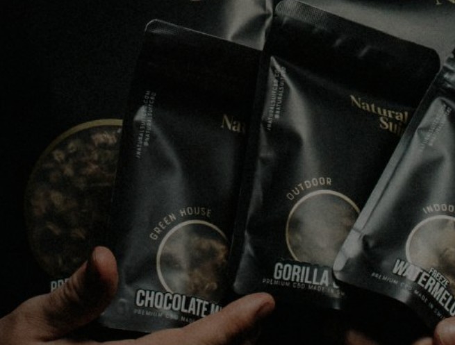 Gros Plan Sur Des Mains Tenant Des Paquets Noirs étiquetés "chocolate Mint", "gorilla" Et "watermelon", Mettant En évidence Les Variétés D'intérieur Et D'extérieur Disponibles Pour L'achat De Cbd En Ligne.