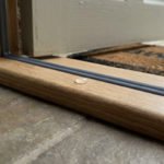 Close-up of a new wooden door threshold with a dark gray sealant and a water bead, highlighting weather protection and quality.