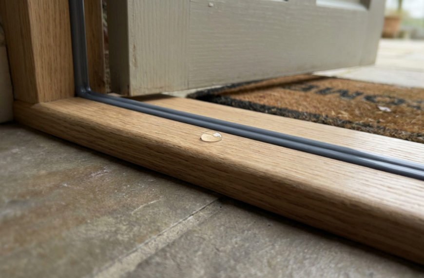Close-up of a new wooden door threshold with a dark gray sealant and a water bead, highlighting weather protection and quality.