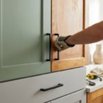 A hand installs a matte black handle on a wooden kitchen cabinet, next to a freshly painted sage green cabinet and grey drawer.