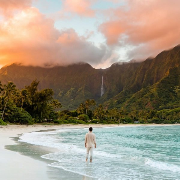 Homme contemplant une plage de sable blanc, mer turquoise et montagnes verdoyantes avec cascade sous ciel orangé.