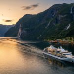 Navire de croisière blanc naviguant dans un fjord norvégien au coucher du soleil. Montagnes verdoyantes, cascades et village côtier.