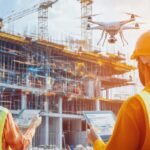 Deux Ouvriers Du Bâtiment En Tenue De Sécurité Utilisent Des Tablettes Tandis Qu'un Drone Vole à Proximité Sur Un Chantier Avec Des échafaudages Et Des Grues En Arrière Plan.
