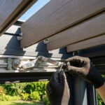 Gros plan sur des mains gantées ajustant le mécanisme d'engrenage de lames orientables en composite pour une pergola, avec jardin en arrière-plan.