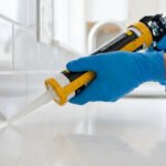 Une personne gantée applique un joint silicone blanc avec un pistolet sur la jonction entre un carrelage mural et un plan de travail.