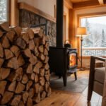 Intérieur chaleureux avec grand tas de bois empilé, poêle à bois allumé, fauteuil confortable et vue sur forêt enneigée.