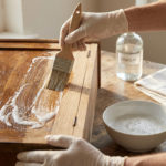 Gros plan sur des mains gantées appliquant une solution moussante avec un pinceau sur un meuble en bois vernis pour le décaper.