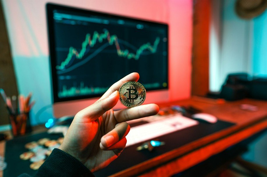 Une Personne Tient Un Jeton Bitcoin Devant Un écran D'ordinateur Affichant Des Graphiques De Cours De Crypto Monnaies Sur Un Bureau.