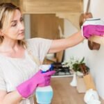 Une Femme Portant Des Gants En Caoutchouc Nettoie Une étagère De Cuisine En Bois à L'aide D'un Chiffon Et D'un Spray Nettoyant. Divers Articles De Cuisine Sont Visibles Sur L'étagère Et Le Plan De Travail.