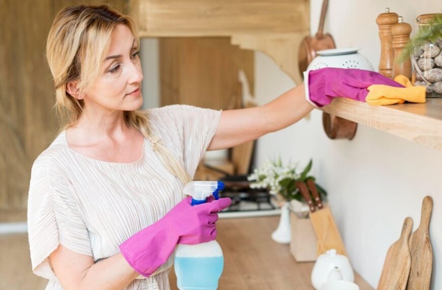 Une Femme Portant Des Gants En Caoutchouc Nettoie Une étagère De Cuisine En Bois à L'aide D'un Chiffon Et D'un Spray Nettoyant. Divers Articles De Cuisine Sont Visibles Sur L'étagère Et Le Plan De Travail.