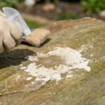 Une main gantée pulvérise un produit pour enlever du ciment séché sur une dalle de pierre, avec une brosse à proximité.