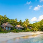 Vue d'une plage de sable blanc avec des bungalows traditionnels aux toits de chaume, bordée de palmiers luxuriants et d'une mer turquoise sous un ciel bleu.