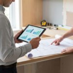 Deux hommes discutent de plans de rénovation sur un comptoir. L'un tient une tablette avec des graphiques, l'autre pointe les plans.