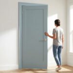 Femme debout face à une porte intérieure bleu-gris dans une pièce lumineuse avec fenêtre, banc et meuble.