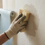 Mains gantées préparant un mur texturé avec une éponge et déroulant un rouleau de papier peint bleu clair, illustrant la rénovation.