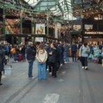 Les Gens Se Promènent Et Font Des Achats Dans Un Marché Couvert Avec Des étals De Nourriture, Des Stands De Produits Frais Et Des Guirlandes Lumineuses.