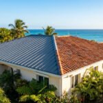 Maison en Martinique avec une toiture neuve en tôle ondulée et une ancienne en tuiles, près de la mer turquoise et des palmiers.