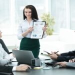 Une Femme Debout Présente Un Tableau à Quatre Collègues Assis Lors D'une Réunion D'affaires Dans Un Bureau Lumineux.