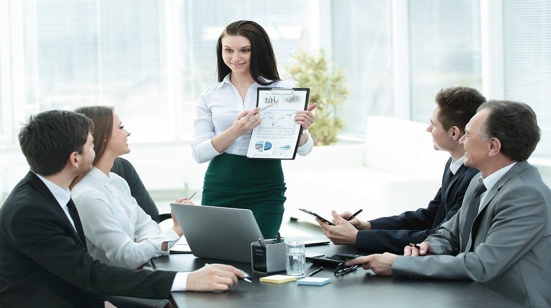 Une Femme Debout Présente Un Tableau à Quatre Collègues Assis Lors D'une Réunion D'affaires Dans Un Bureau Lumineux.